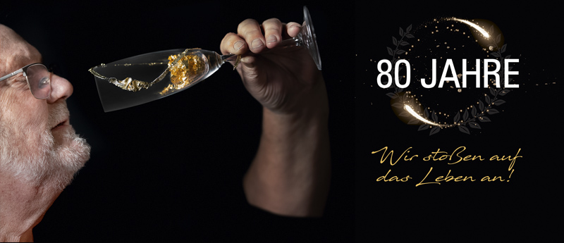Rolf Wingerath stößt mit einem Glas Sekt an, daneben der Schriftzug „80 Jahre – Wir stoßen auf das Leben an!“